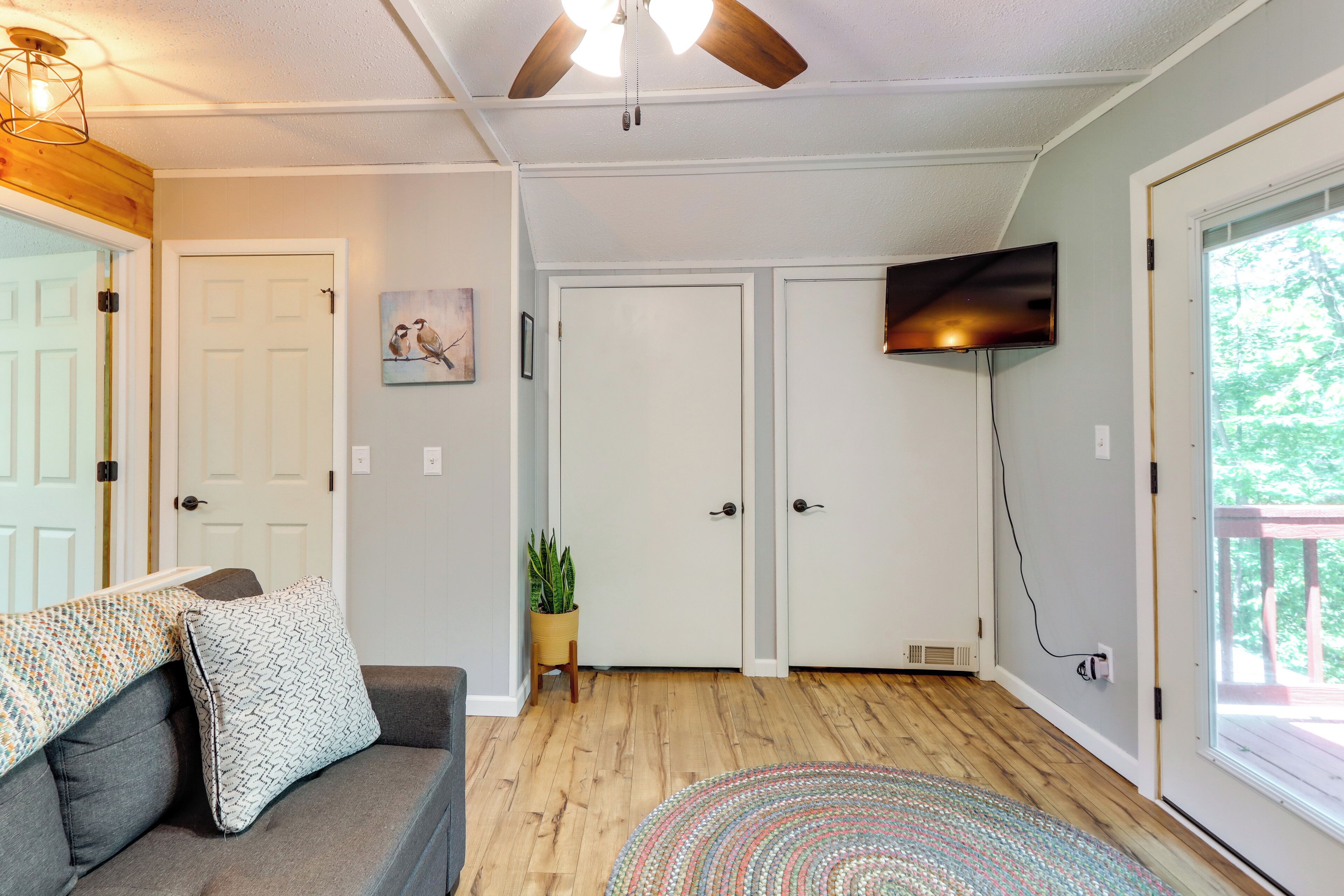 A room with a ceiling fan, a TV mounted on the wall, a wooden floor, a sofa with patterned cushions, and a door with a small plant beside it.