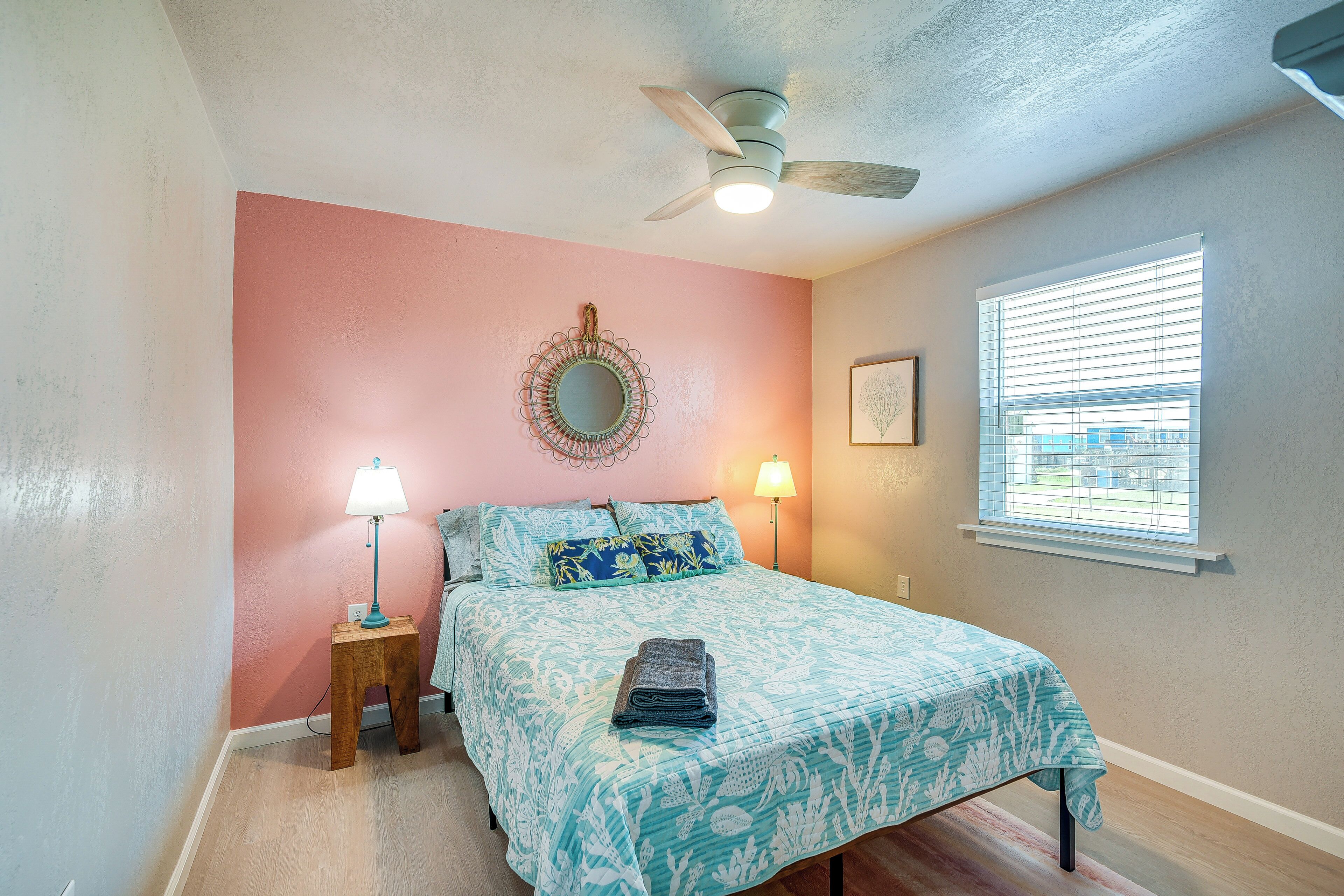 A bedroom with a bed, a ceiling fan, a window with blinds, a bedside lamp, and a wall mirror.