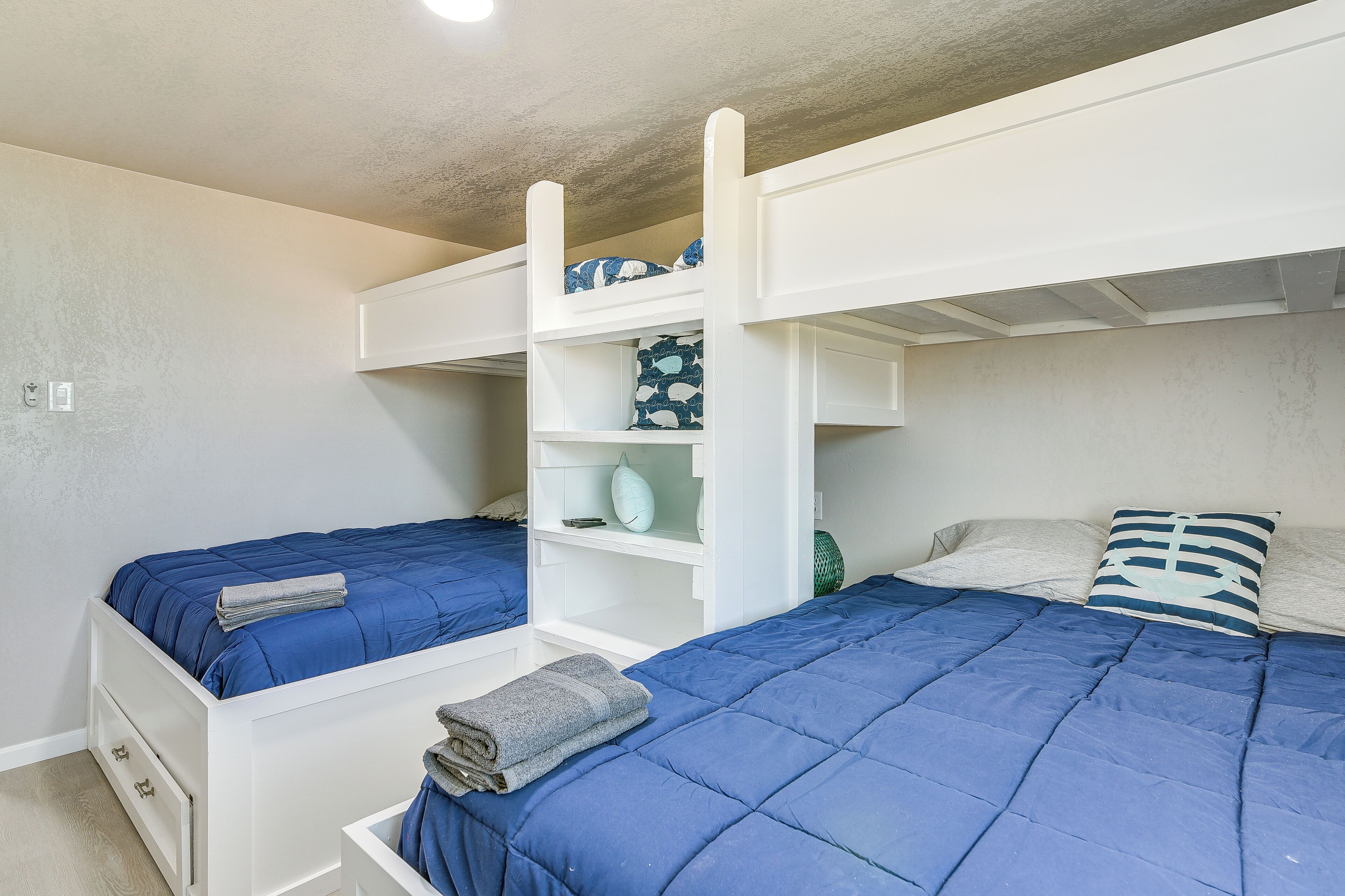 Une chambre avec un lit superposé à la structure blanche, deux lits avec une literie bleue, une étagère avec des objets décoratifs et un petit tiroir.