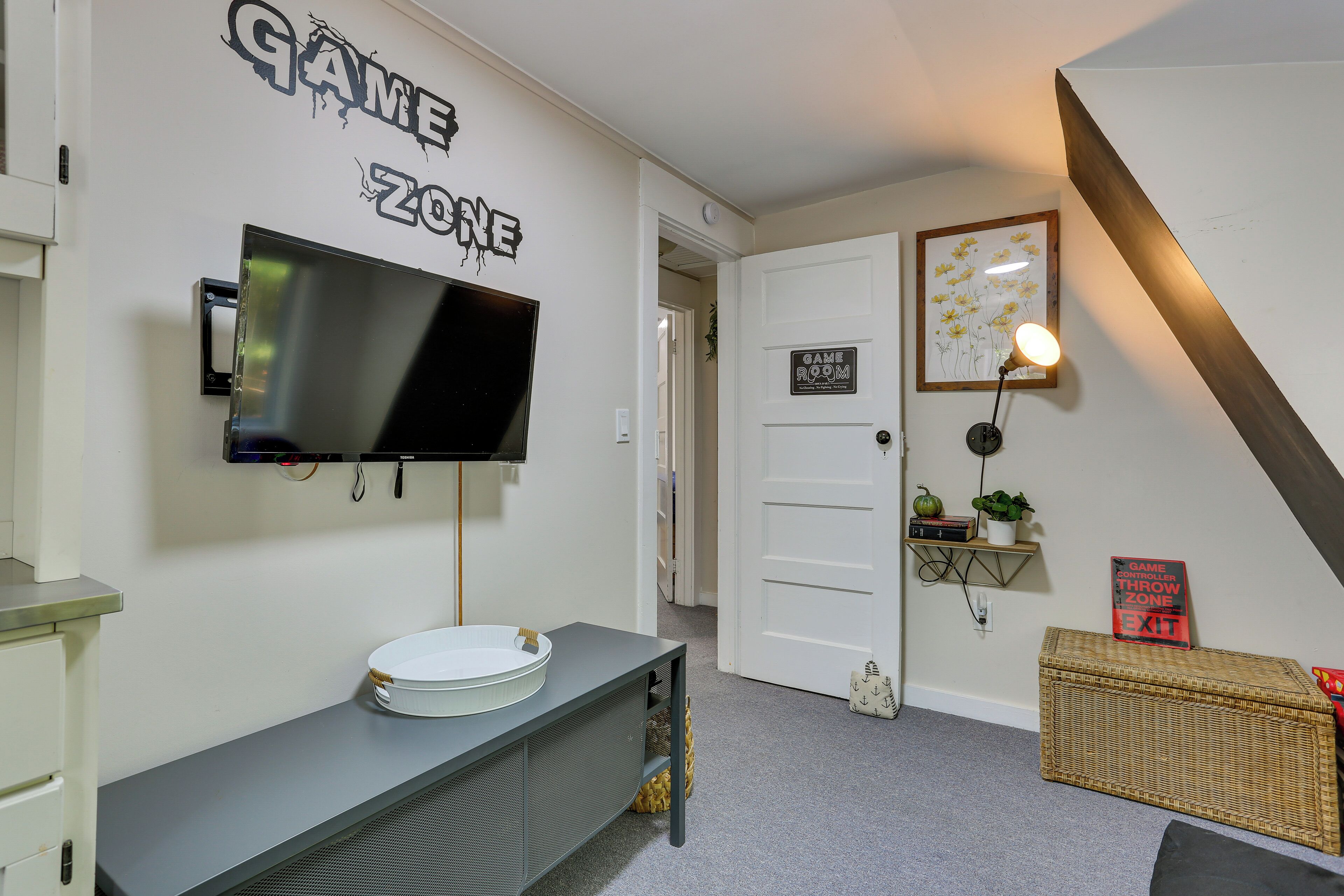 A room with a mounted TV, a white door, a Game Zone sign, a wicker chest, and a framed picture on the wall.