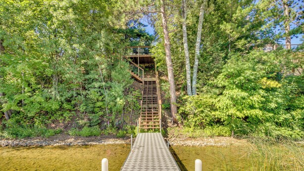 A wooden deck leading to a wooden staircase surrounded by dense greenery. - Lakefront Minnesota Escape w/ Fire Pit & Boat Dock (Emily)