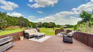 A wooden deck with outdoor furniture, including a sofa, chairs, and a table, overlooking a grassy yard with trees and a distant building. - Inviting Granbury Home w/ Fire Pit: 3 Mi to Lake! (Granbury)