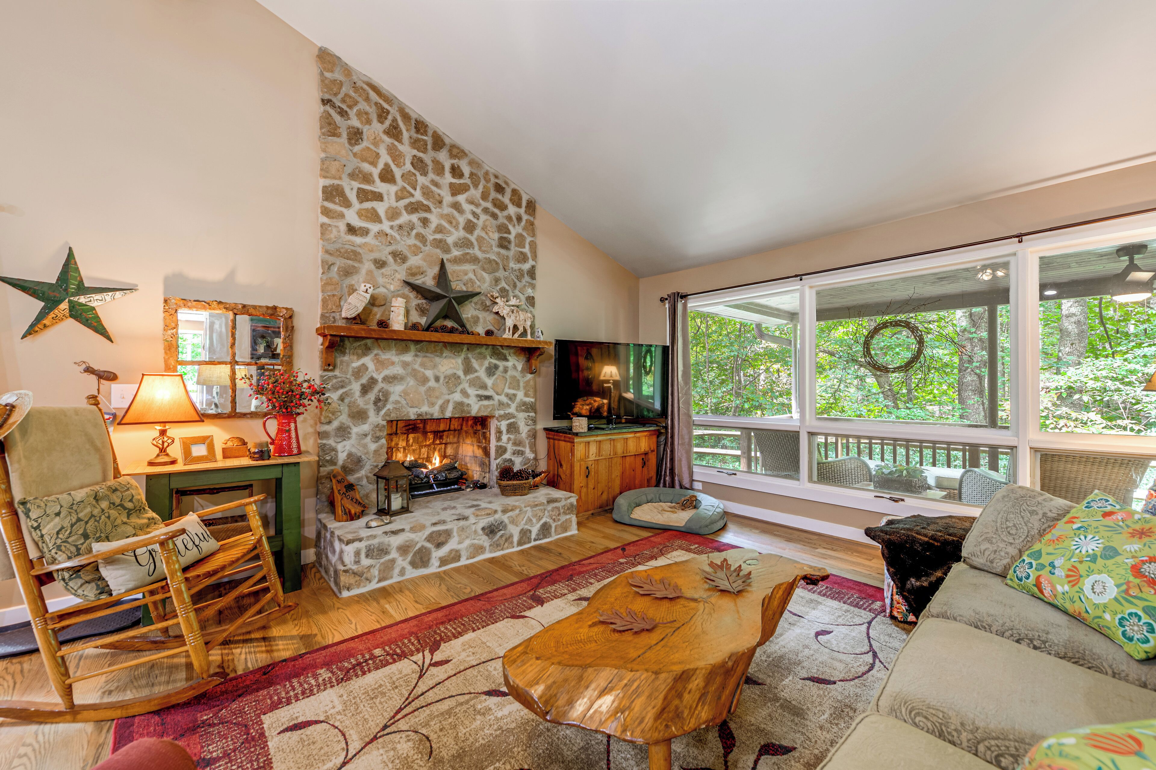 A cozy living room with a stone fireplace, a large window with a view of greenery, a rocking chair, and a sofa.