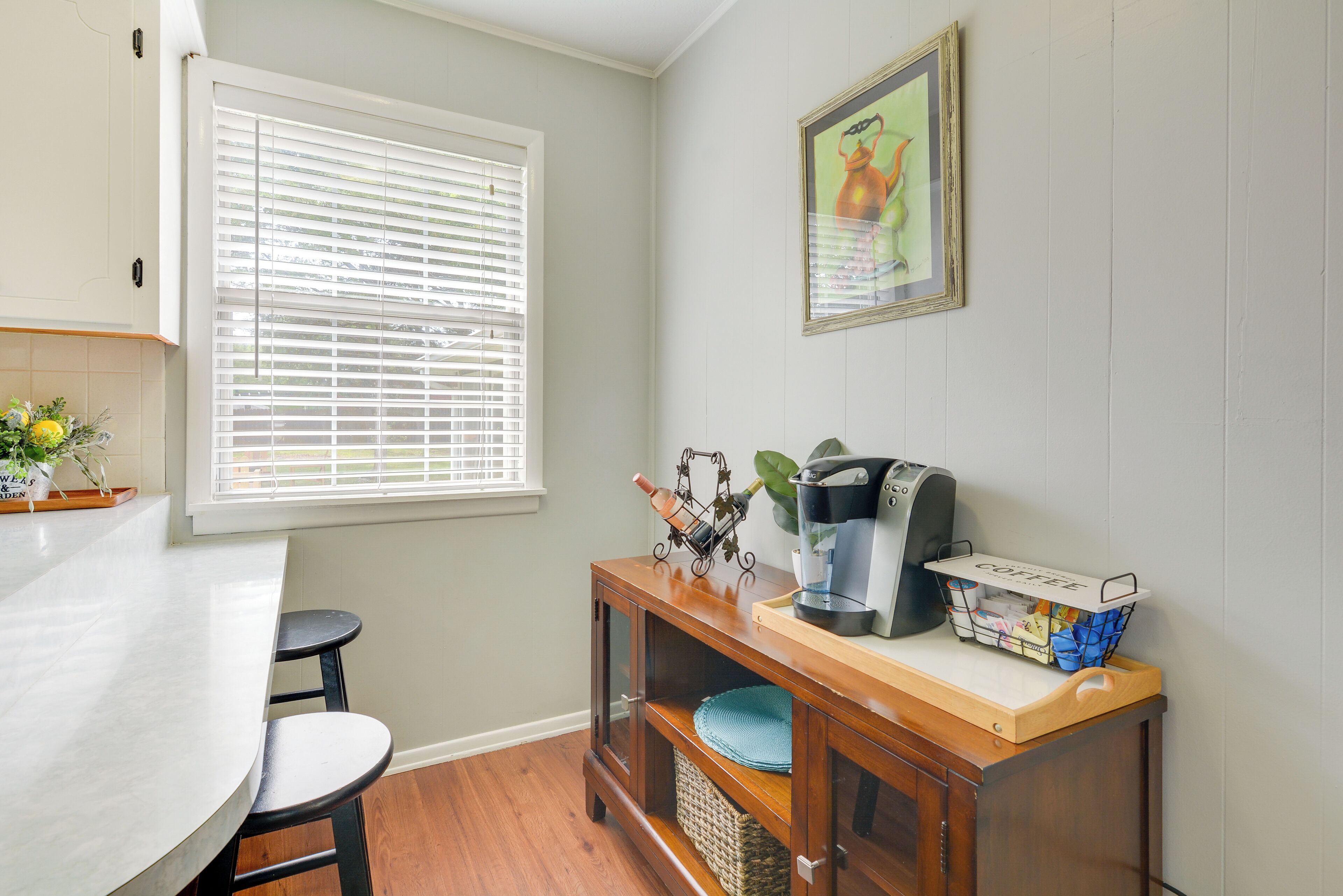 A kitchen with a white countertop, a wooden cabinet, a coffee machine, and a basket of coffee pods.