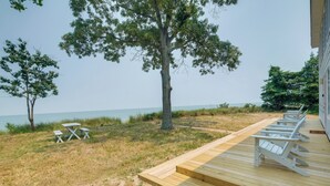 Une terrasse en bois avec des chaises blanches et une petite table, donnant sur une étendue d’eau, avec des arbres en arrière-plan.