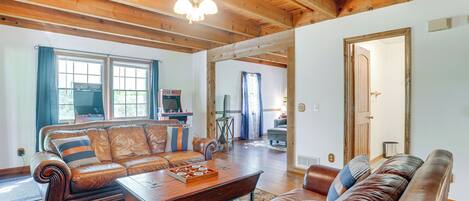 A spacious living room with a wooden ceiling and a large ceiling fan, a brown leather sofa, a wooden coffee table, and a view of the adjacent kitchen through an open doorway.