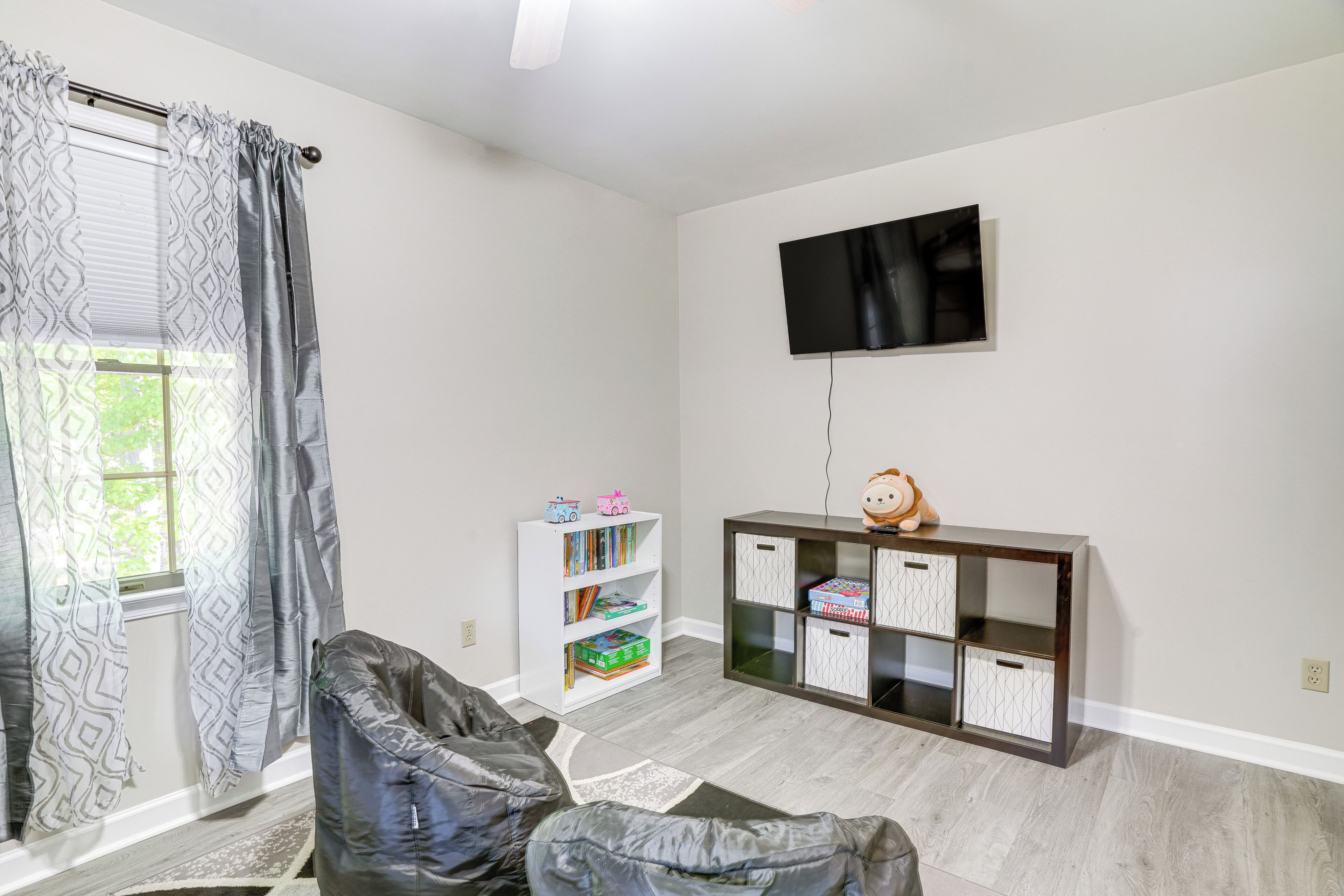 Ein Zimmer mit großem Fenster, einem flachbildfernseher an der Wand, einem Bücherregal und einer Kommode mit Schubladen.