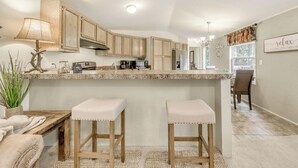 Eine Küche mit Holdelementen, einer Granit Arbeitsplatte, einer Mikrowelle und einem Kühlschrank. Zwei Barhocker und ein Tisch mit einer Lampe sind vorhanden.