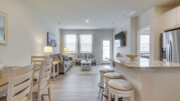 A modern kitchen with a central island, bar stools, and a dining area with wooden chairs. - Shuttle to Beach: Millville By the Sea Retreat (Millville)