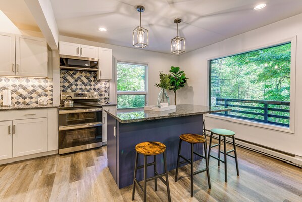 A modern kitchen with a large island, stainless steel appliances, and a view of a green landscape through large windows. - Waynesville Cabin w/ Pickleball Court and Hot Tub (Waynesville)