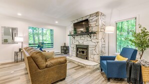 A modern living room with a stone fireplace, a large flat-screen TV, a blue armchair, and a brown sofa. - Waynesville Cabin w/ Pickleball Court and Hot Tub (Waynesville)
