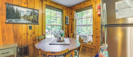 A wooden interior with a dining table, chairs, a refrigerator, and a window with a view of trees.