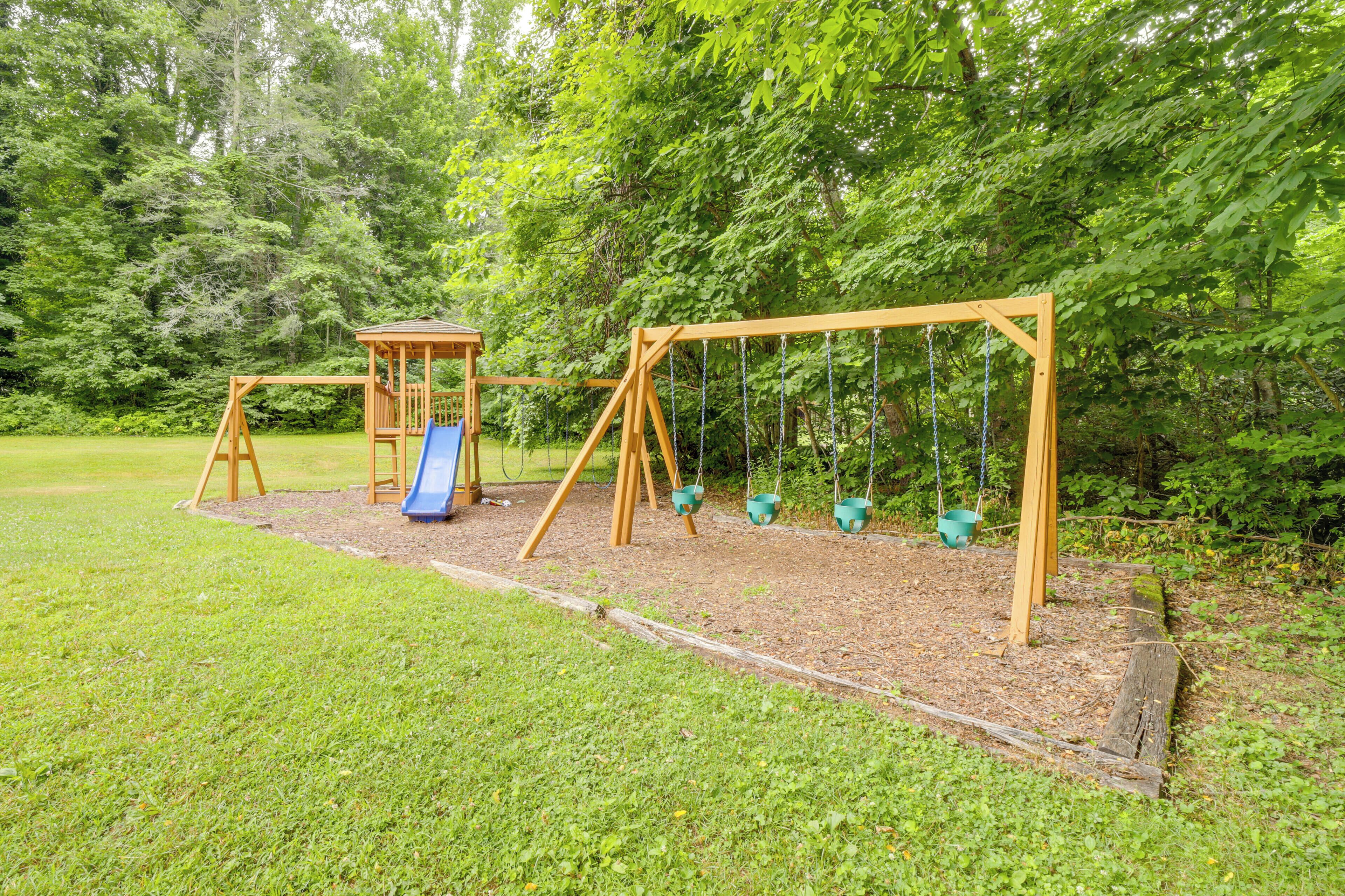 Un parque infantil con tobogán y columpios, rodeado de árboles y césped.