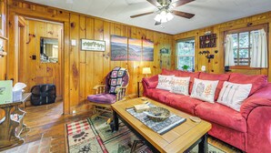 A cozy cabin interior with wooden walls, a red sofa, a wooden coffee table, and a ceiling fan. - Pet-friendly Creekside Cabin w/ Pool Access! (Maggie Valley)