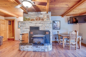 A stone fireplace with a television above it, wooden dining table and chairs, and a ceiling fan.