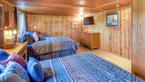 Ein Zimmer mit Holzwandverkleidung, zwei Betten, einem Schminktisch, einem Fernseher und einem gerahmten Bild an der Wand.