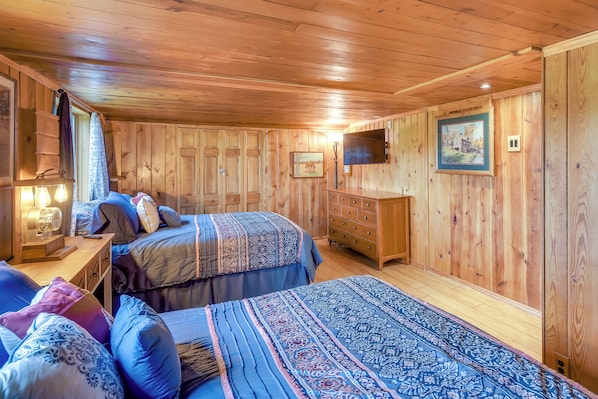 Quarto com painéis de madeira, duas camas, cômoda, televisão e um quadro na parede.