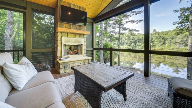 A cozy outdoor seating area with a stone fireplace, a flat-screen TV, and a view of a lake surrounded by trees. - Grill + Water Views: Chinquapin Resort Home (Glenville)