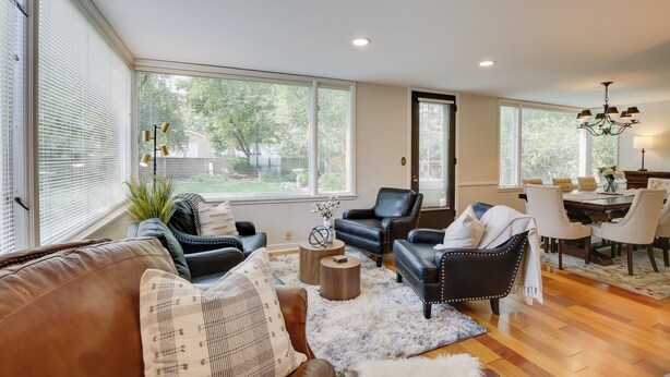 Un salon spacieux avec un canapé en cuir, des fauteuils en cuir noir, une table à manger avec des chaises blanches et de grandes fenêtres équipées de stores.