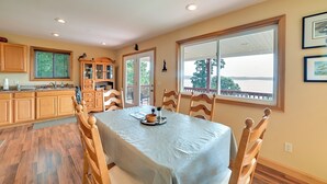A dining area with a table set for four, wooden chairs, a kitchen with wooden cabinets, and a view through a large window. - Deck & Water View: Cozy Kentucky Lake Getaway (New Concord)