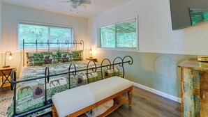 Quarto com cama, um banquinho, ventilador de teto e duas janelas com persianas.
