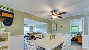 Uma sala de jantar com uma mesa e cadeiras brancas, um ventilador de teto e vista para a sala de estar.