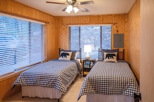 A cabin bedroom with two beds, wooden walls, a ceiling fan, and a window with blinds.