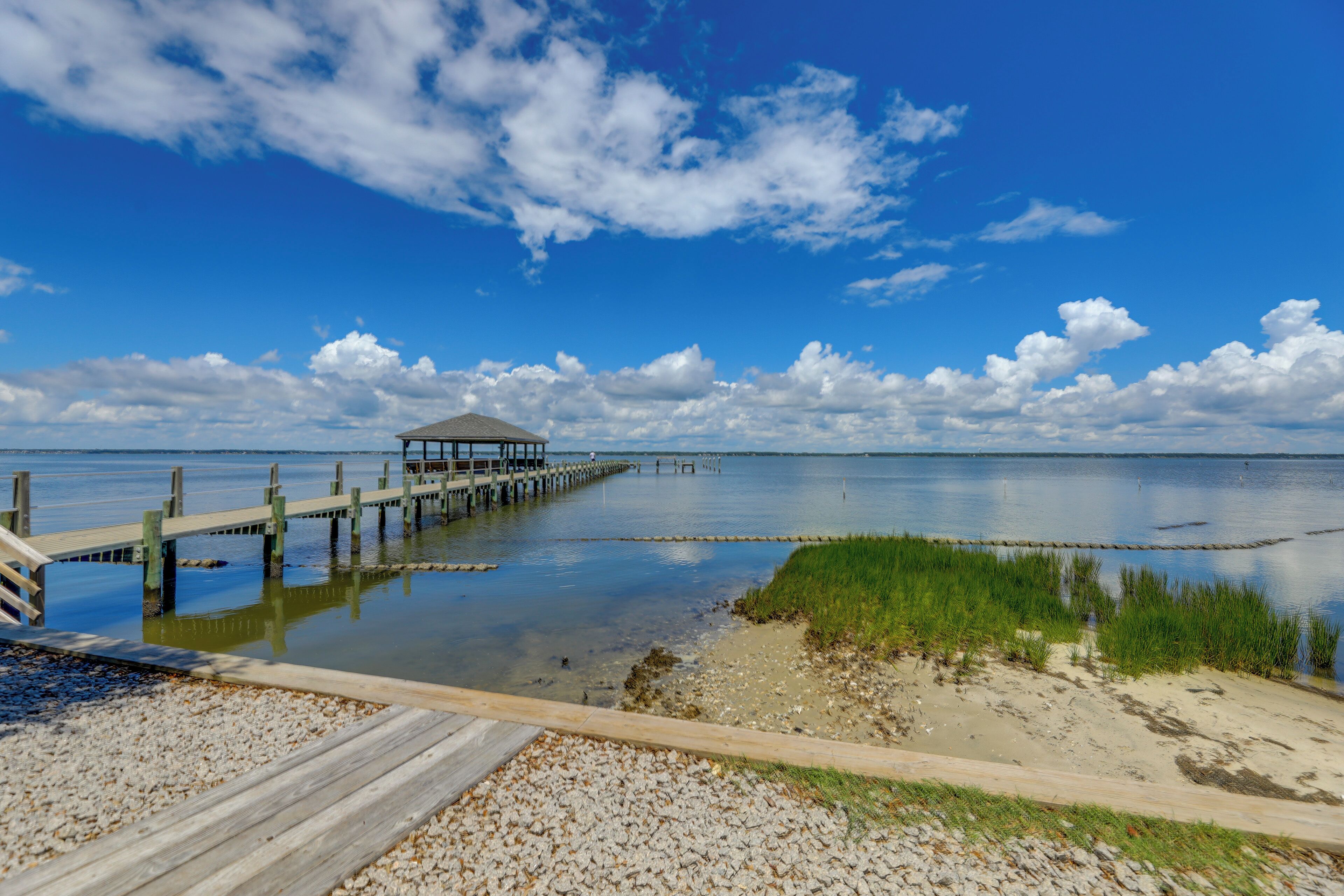 A wooden pier extending into calm waters, a grassy area, and a clear blue sky with clouds.