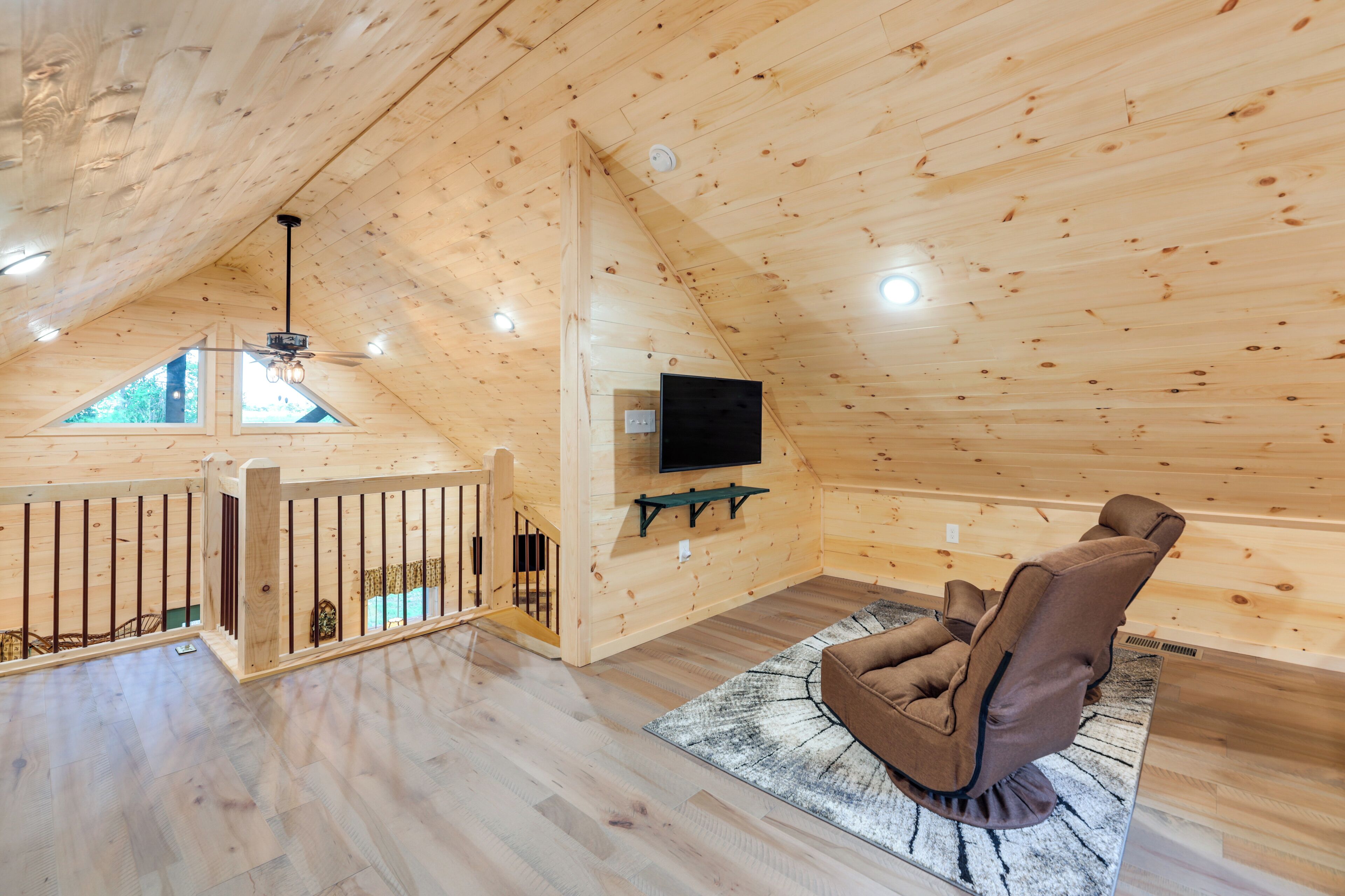 Un interior de madera con techo inclinado, un televisor montado, un sillón reclinable marrón y vistas a través de una ventana.