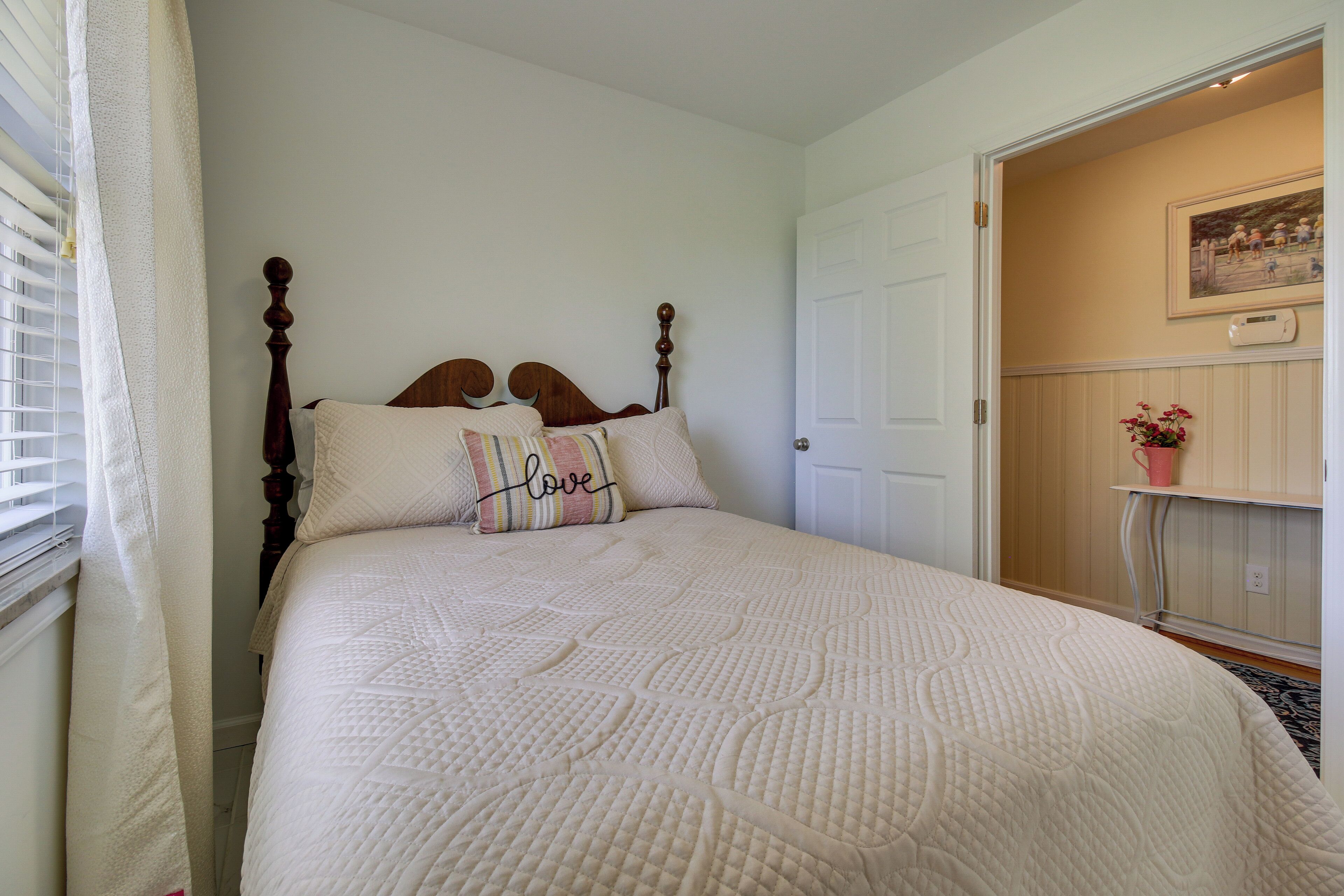 A bedroom with a bed, a door, a window with blinds, and a small table with a flower vase.