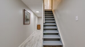 Un couloir étroit avec un plancher en bois, un escalier aux marches blanches et un tableau encadré accroché au mur.