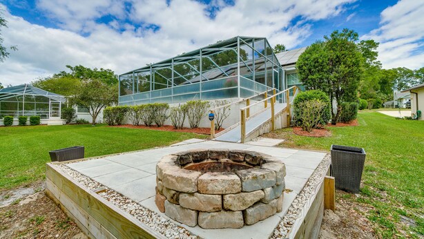 A modern outdoor patio area with a stone fire pit, a glass greenhouse structure, and a well-maintained lawn.