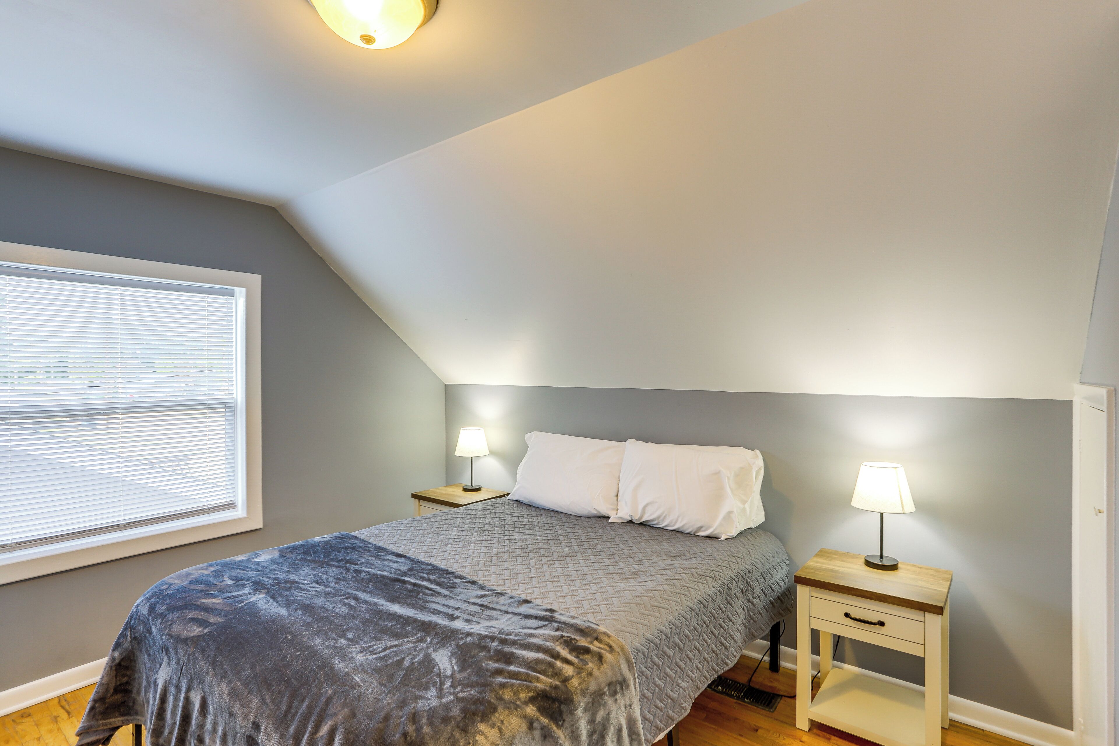 A bedroom with a bed, two bedside tables with lamps, a window with blinds, and a sloped ceiling.