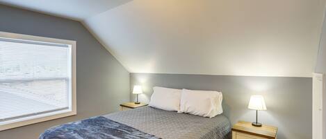 A bedroom with a bed, two bedside tables with lamps, a window with blinds, and a sloped ceiling.