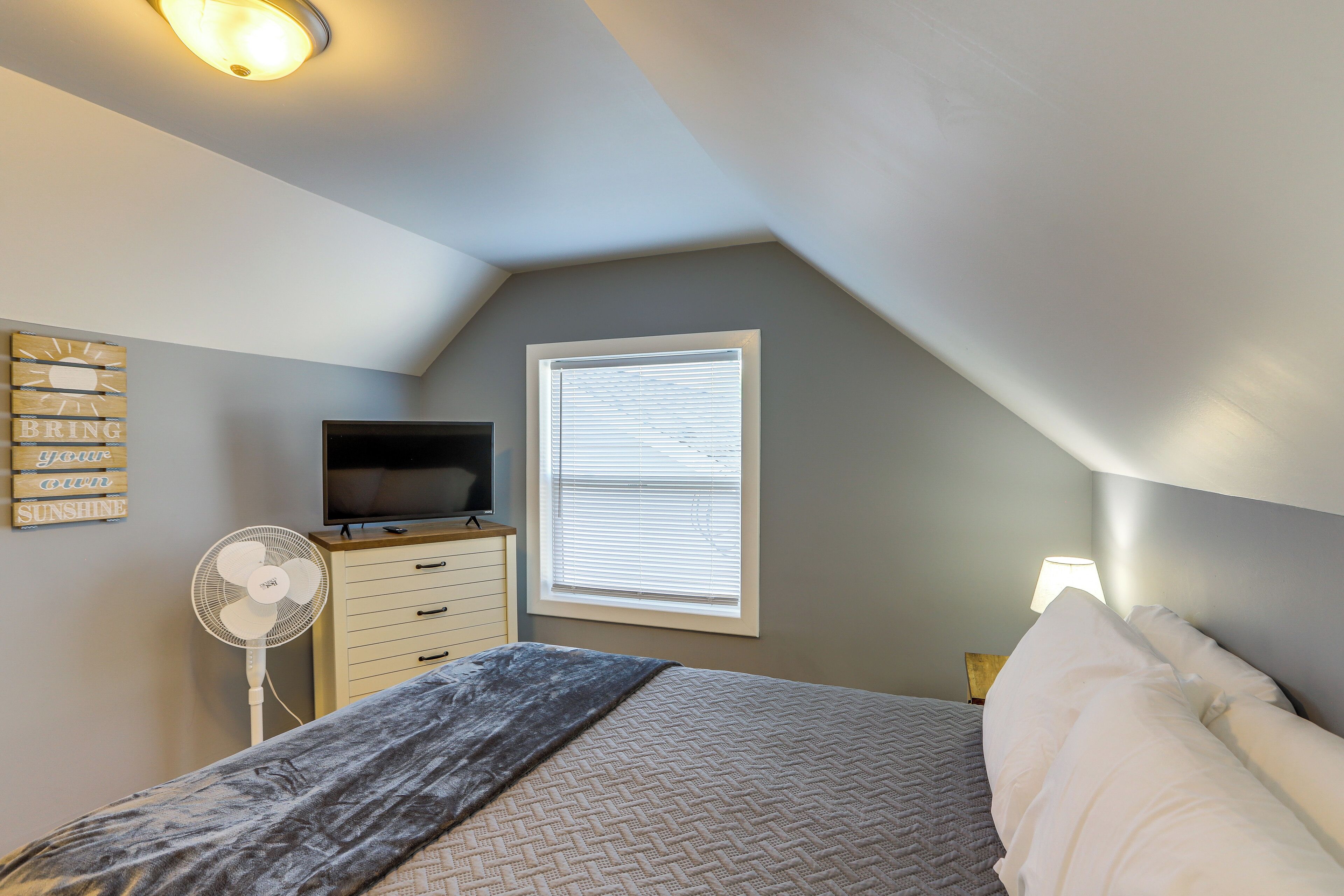 A bedroom with a bed, a TV, a fan, a window with blinds, and a wall-mounted light.