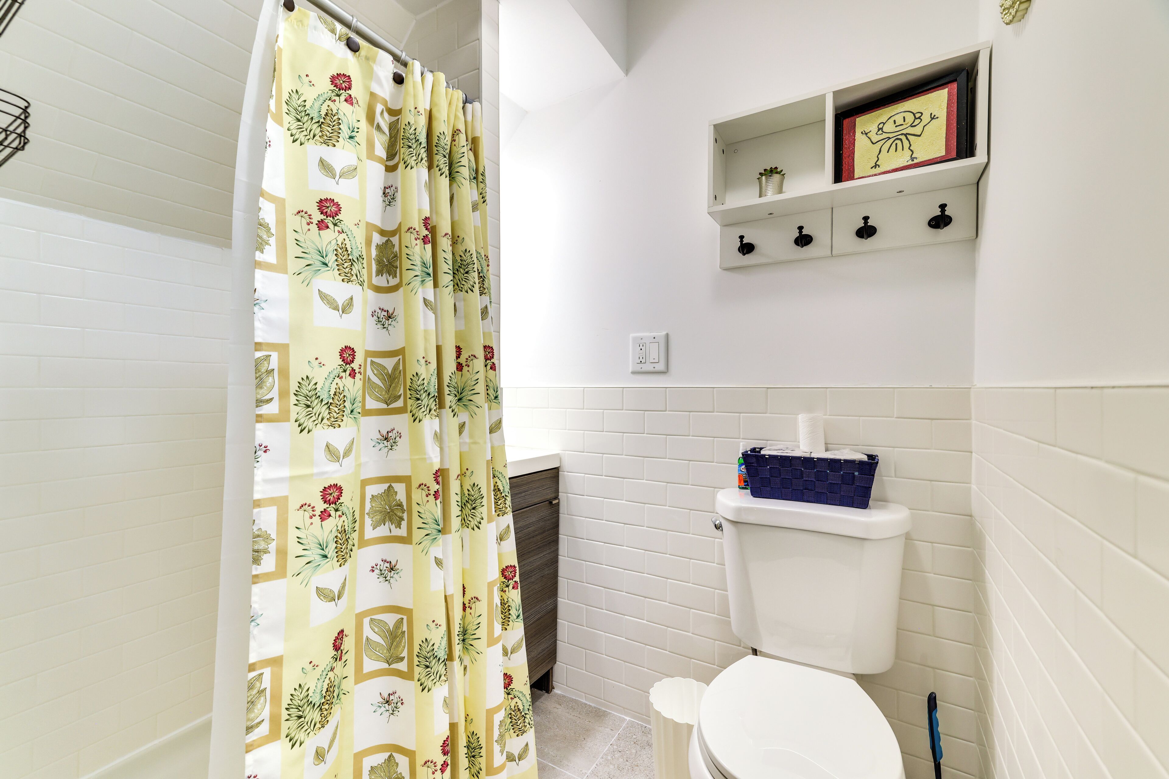 Une salle de bain comprenant des toilettes, un rideau de douche, une étagère avec des articles de toilette et un petit tableau encadré.