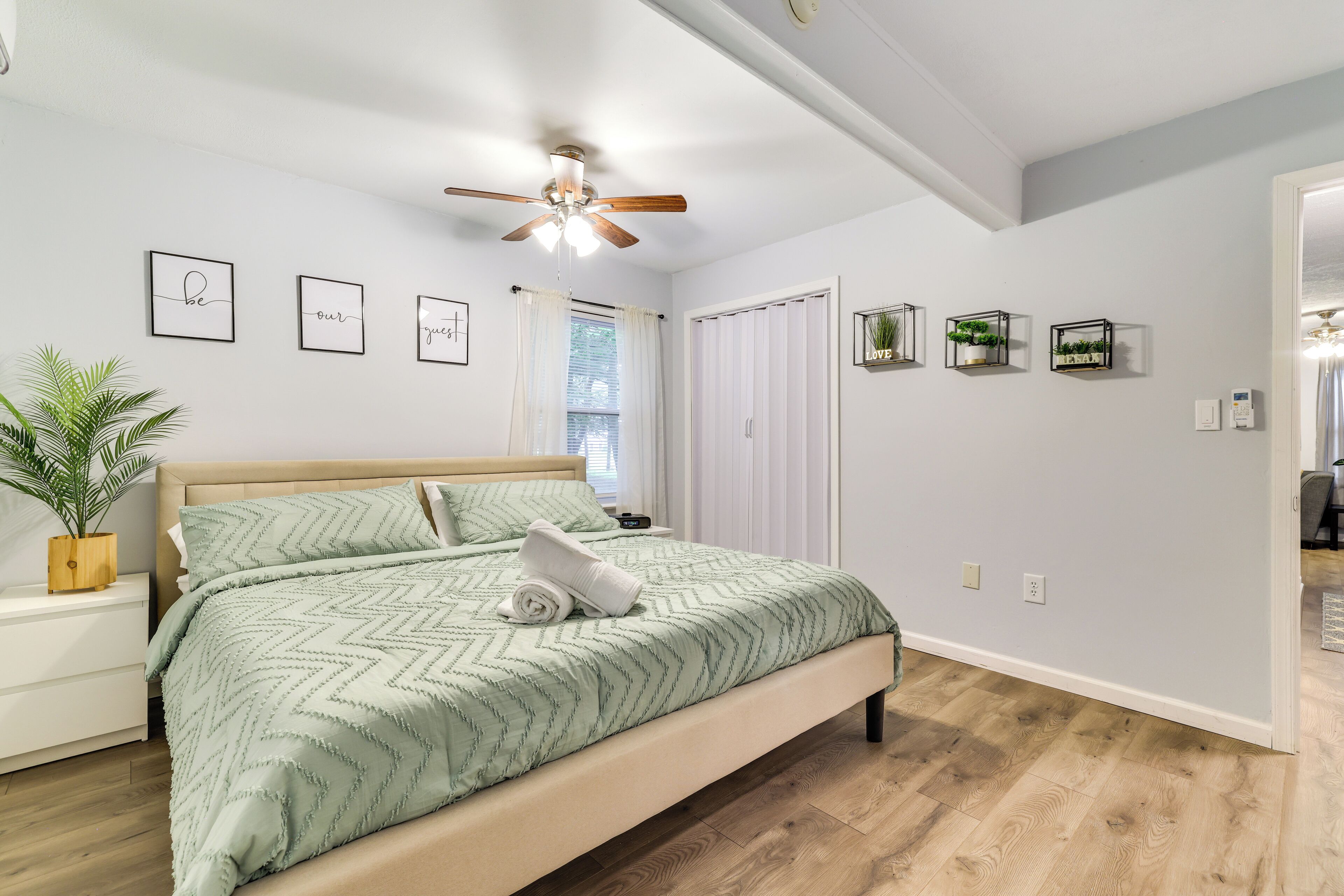 A bedroom with a bed, a ceiling fan, a bedside table with a plant, and framed pictures on the wall.