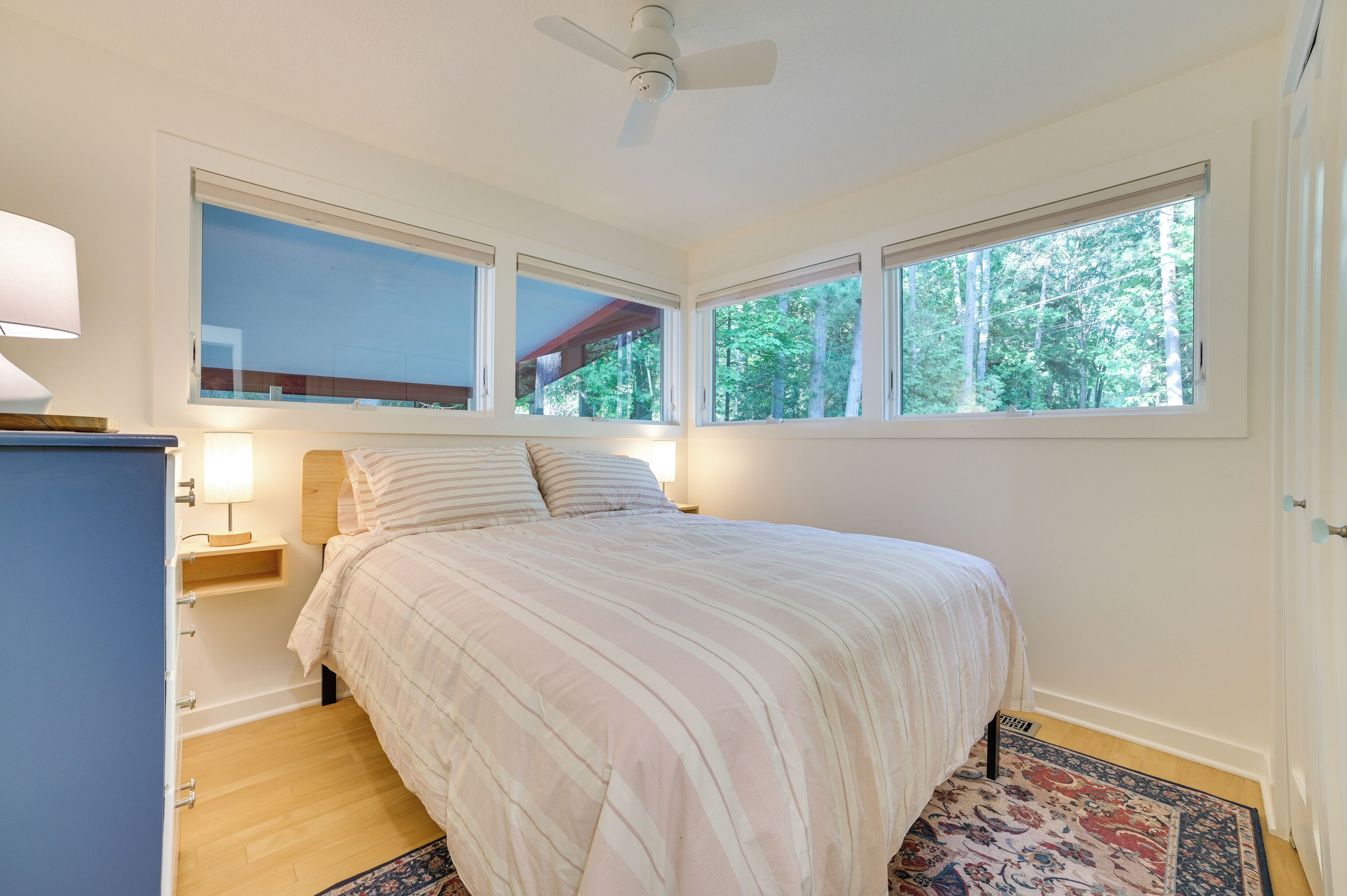 Quarto com cama, mesas de cabeceira, ventilador de teto e grandes janelas com vista para árvores.