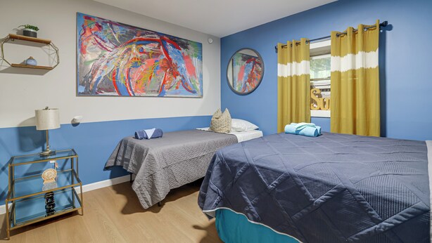 Un dormitorio con dos camas, un cuadro abstracto grande, un espejo redondo y una ventana con cortinas amarillas.