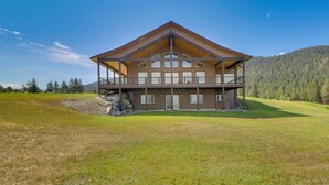 A large, two-story building with a prominent wooden roof and multiple windows, situated on a grassy field with a forested mountain in the background.