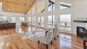 A spacious living room with a high ceiling, large windows, a fireplace, and a dining area.