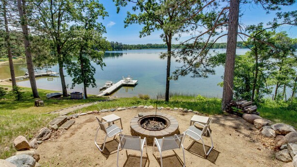 A lakeside outdoor seating area with a fire pit, surrounded by rocks and trees, overlooking a calm lake with a dock and boats. - Waterfront Pequot Lakes Cabin: Fire Pit + Views! (Pequot Lakes)