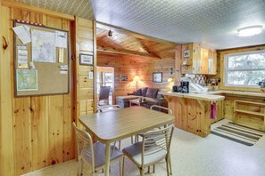 A cozy cabin-style interior with a wooden dining area, a kitchen with a large window, and a living space with a sofa and lamp. - Waterfront Pequot Lakes Cabin: Fire Pit + Views! (Pequot Lakes)