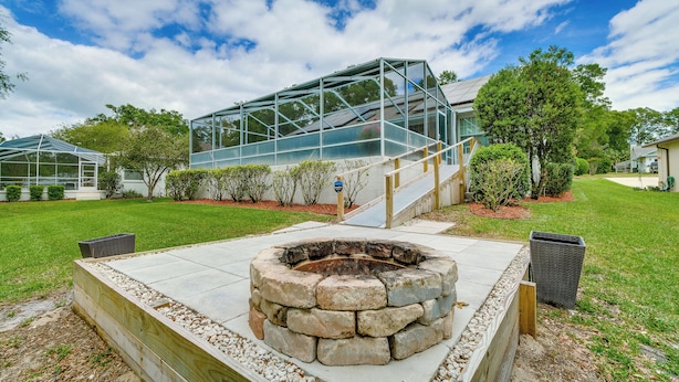A modern outdoor patio area with a stone fire pit, a glass greenhouse structure, and a well-maintained lawn. - Lanai & Grill: Sunny Home Near Rainbow Springs! (Dunnellon)