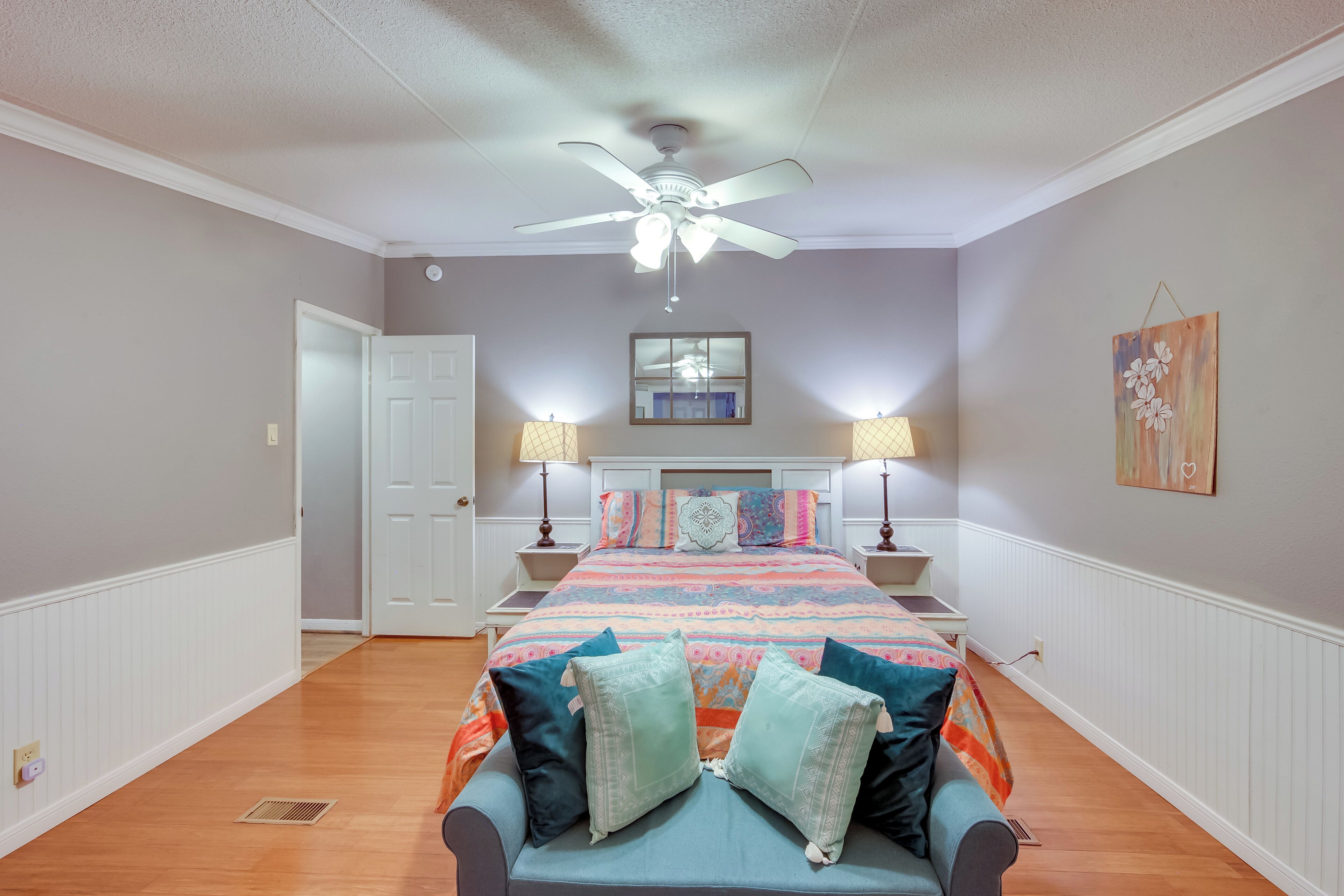 A bedroom with a bed, two bedside lamps, a ceiling fan, a door, and a wall-mounted artwork.