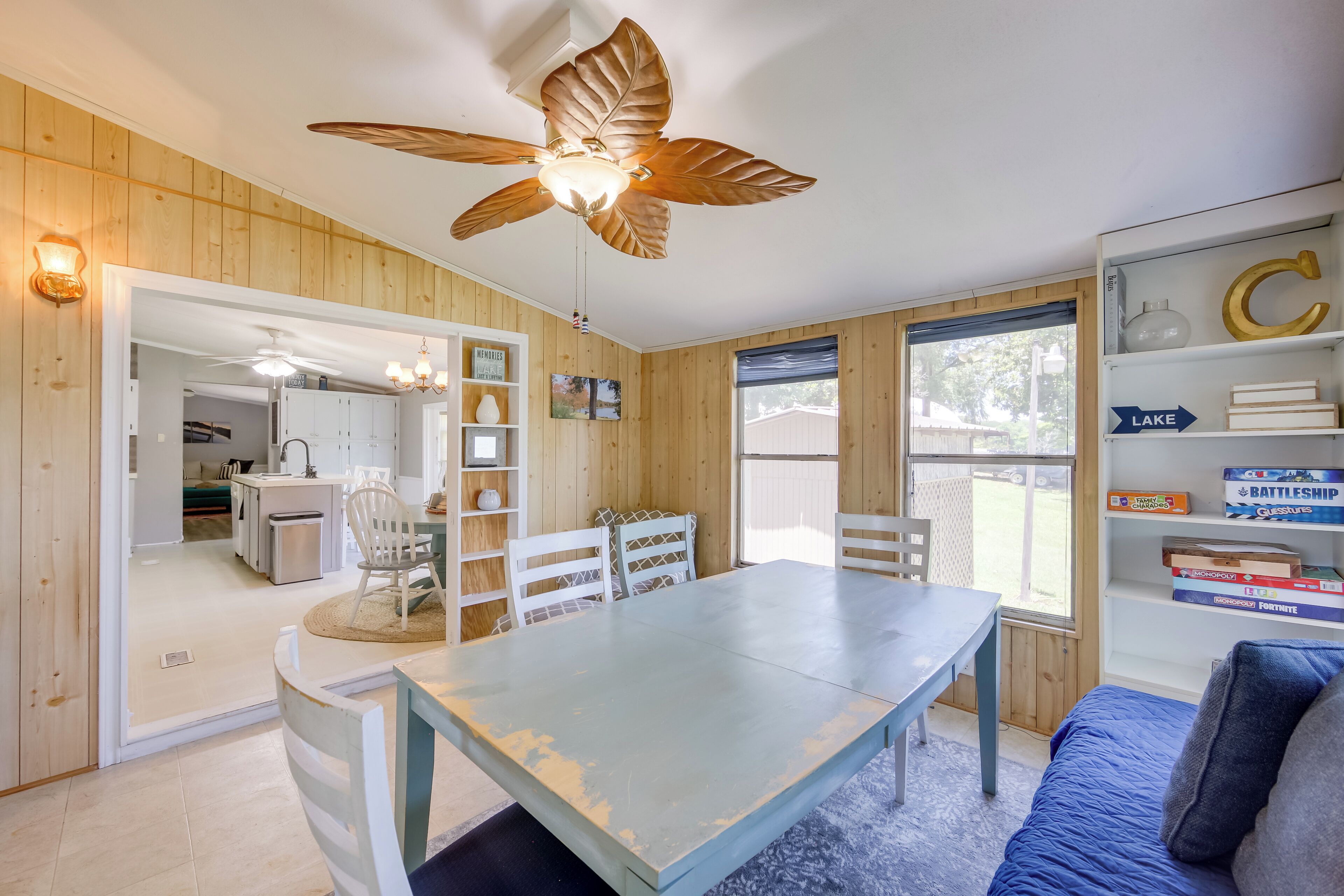 Una habitación con ventilador de techo de madera, una mesa de comedor con sillas, una estantería y una ventana con vistas al exterior.