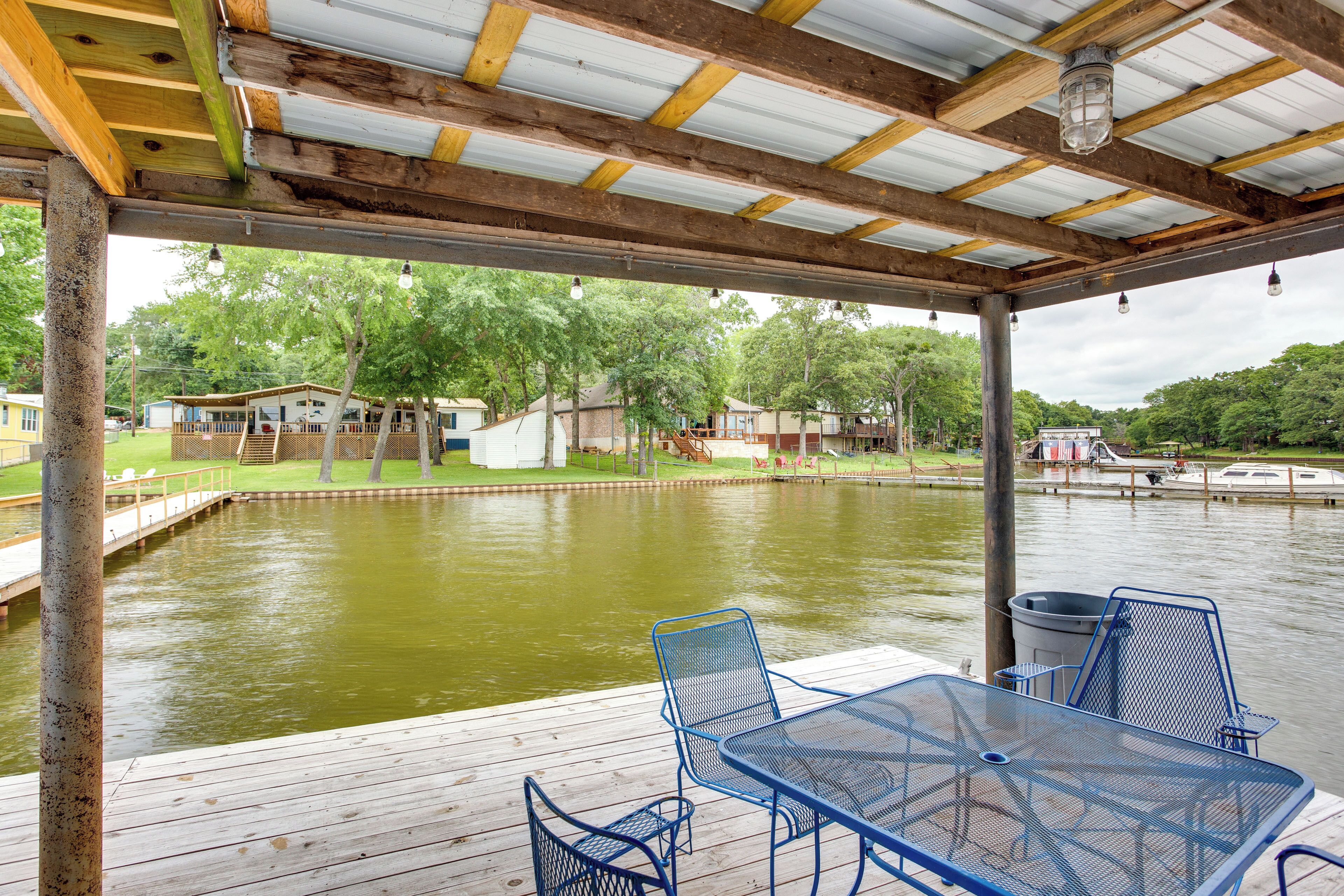 A covered outdoor area with a wooden deck, metal chairs, and a table overlooking a calm body of water with boats in the background.