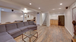 Een moderne woonkamer met een grijze hoekbank, een glazen salontafel en een houten vloer. Er is een keuken met een koelkast en houten kasten.