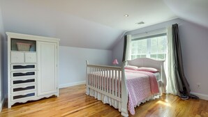 A bedroom with a crib, a white cabinet with drawers, a window with curtains, and a wooden floor. - Spacious Connecticut Home - Deck, Grill & Fire Pit (Mystic)
