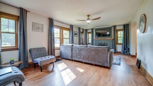 A spacious living room with a large grey sofa, a grey armchair, and a small table with a plant. There is a ceiling fan, a fireplace, and a large window with curtains. - Central Erie Vacation Rental ~ 2 Mi to Beach! (Erie)